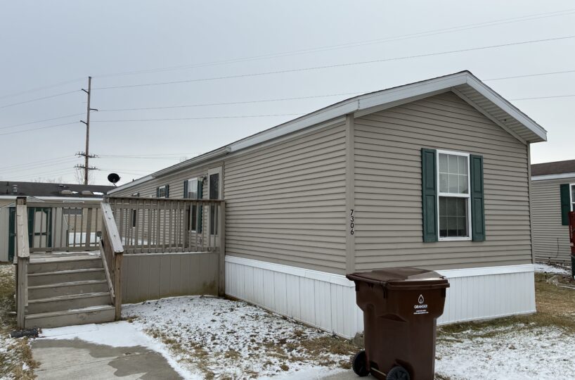 Beige manufactured home with green shutters, a wooden deck and stairs, and a brown trash bin in a snowy yard.