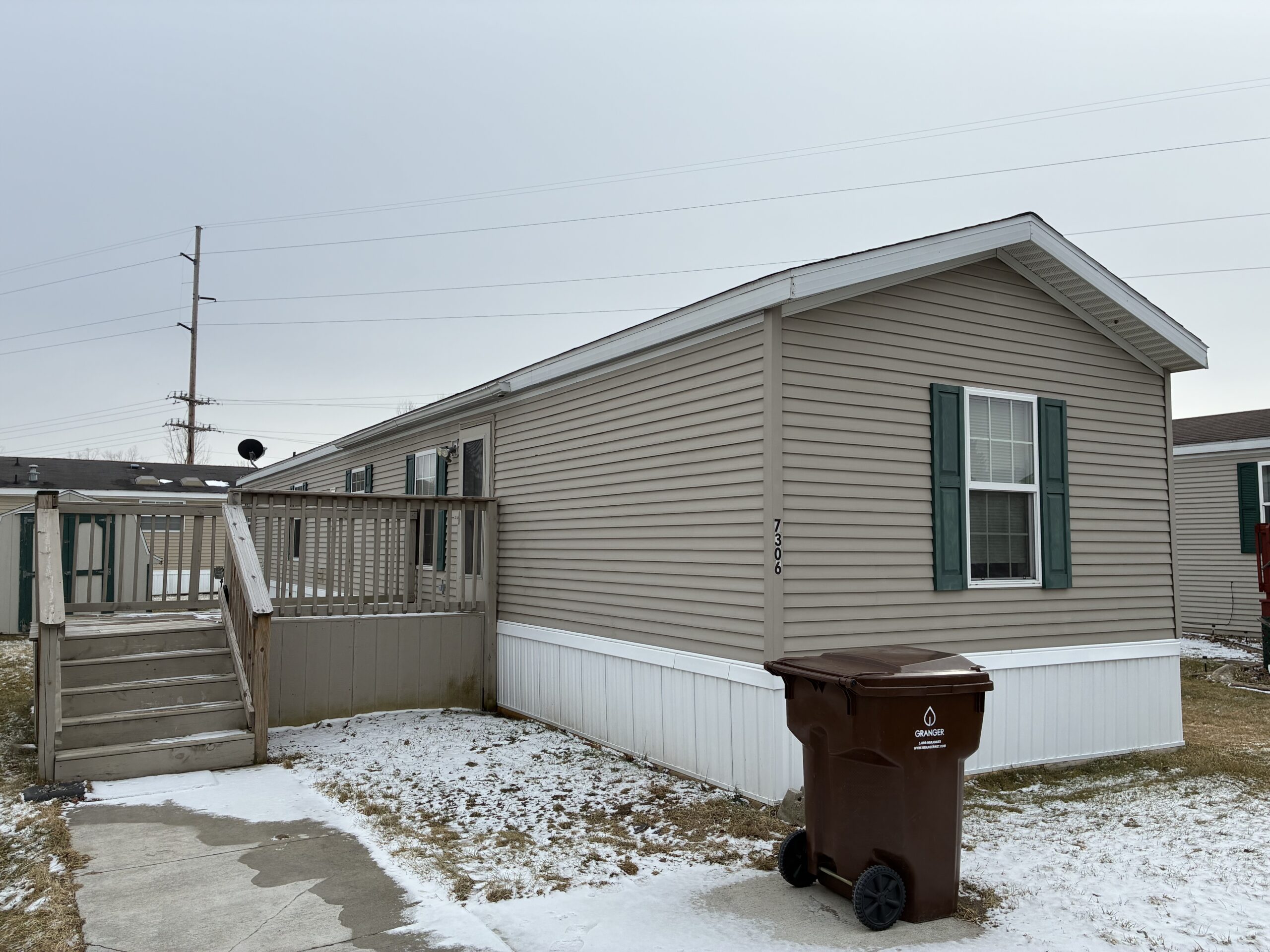 Beige manufactured home with green shutters, a wooden deck and stairs, and a brown trash bin in a snowy yard.